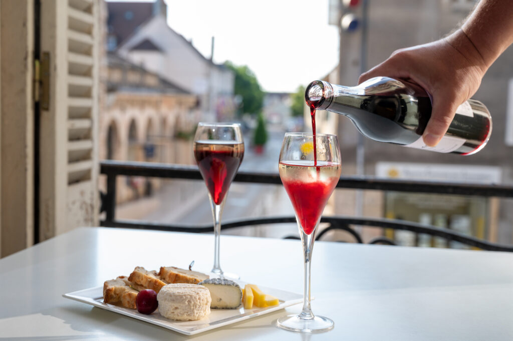 Drinking of Kir Royal, French aperitif cocktail made from creme de cassis topped with champagne, typically served in flute glasses, with view on old French village