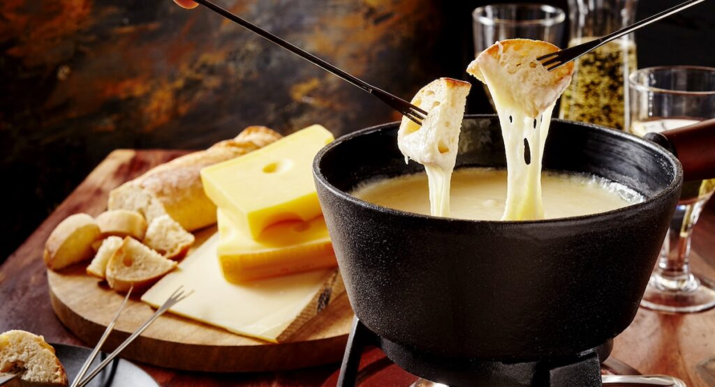 Gourmet Swiss fondue dinner on a winter evening with assorted cheeses on a board alongside a heated pot of cheese fondue with two forks dipping bread and white wine behind in a tavern or restaurant