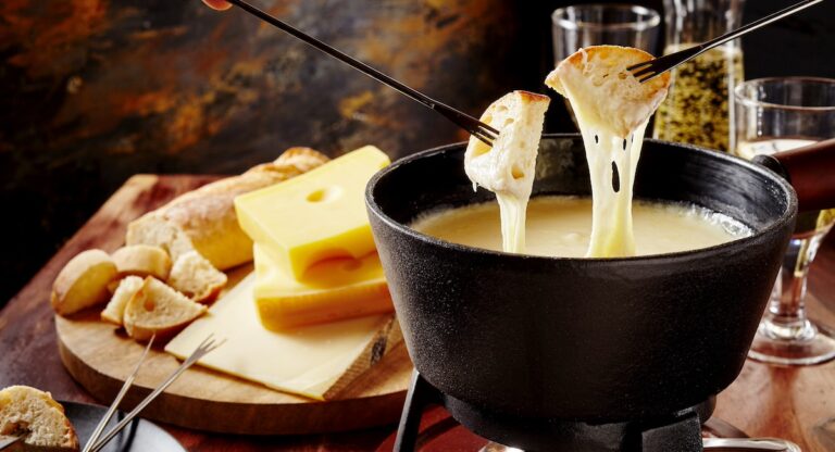 Gourmet Swiss fondue dinner on a winter evening with assorted cheeses on a board alongside a heated pot of cheese fondue with two forks dipping bread and white wine behind in a tavern or restaurant