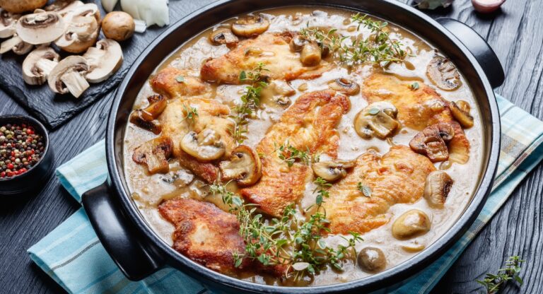 Chicken Marsala, Italian-American dish of golden pan-fried chicken cutlets and mushrooms in a rich Marsala wine sauce in a black ceramic dish on a wooden table, view from above, close-up