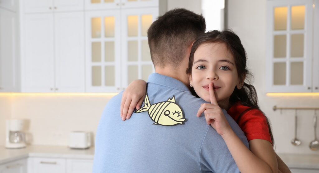 Cute little girl sticking paper fish to father's back at home