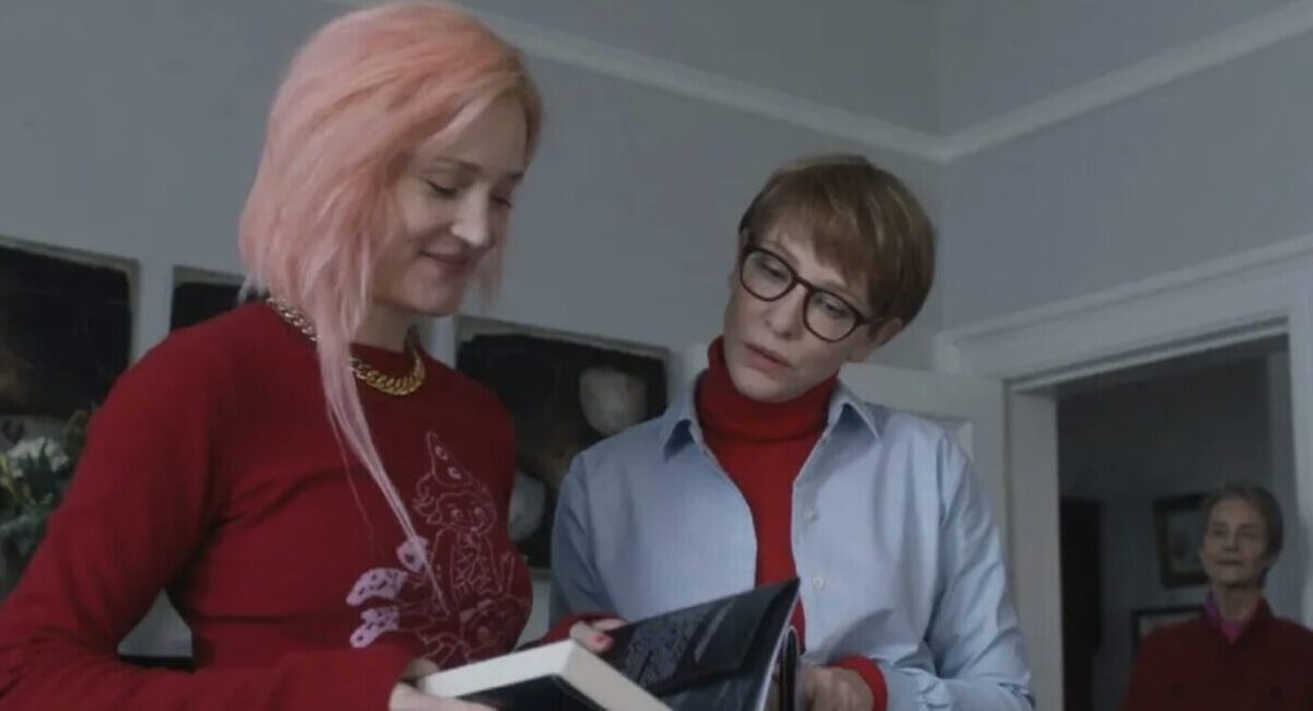 Woman with pink hair reading book and smiling beside older woman wearing glasses.