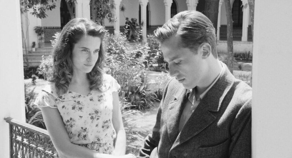 A man looks down while a woman smiles at him in a black and white film still.