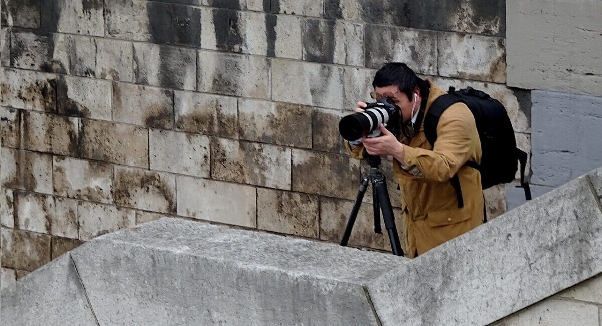 Man wearing backpack pointing film camera with stone wall backdrop.