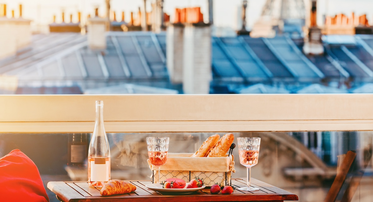 Pink wine in two glasses, traditional french bakery products - baguettes, macaron, croissant and strawberries on a balcony with a view on rooftops and Eiffel Tower