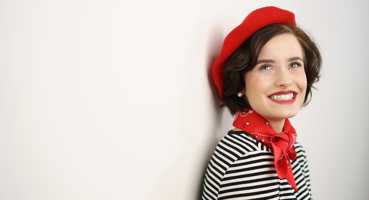 Attractive elegant young woman in French fashion wearing a bright red beret and scarf standing leaning back against a wall looking up with a happy smile