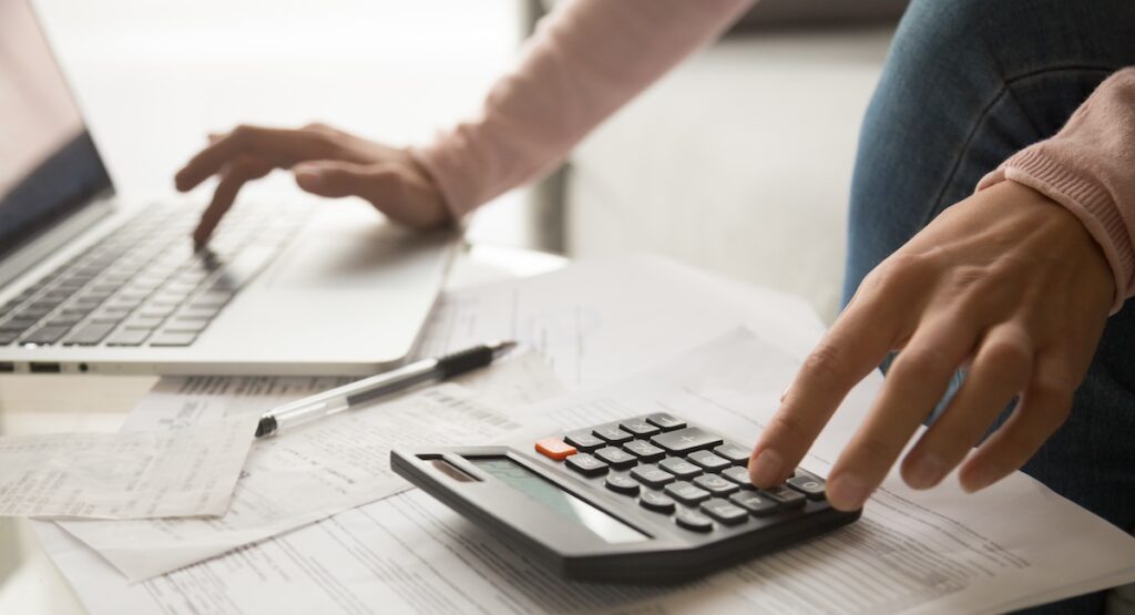 Close up cropped image young woman calculating monthly expenses, managing budget, entering data in computer application, sitting at table full of papers, loan documents, invoices, utility bills.