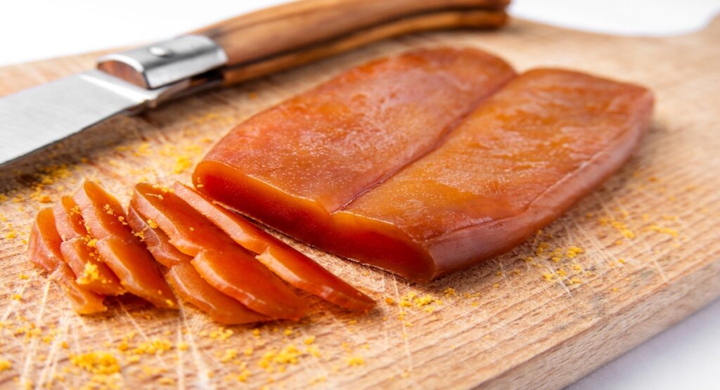 A picture of sliced poutargue on a wooden cutting board next to a wooden-handled knife