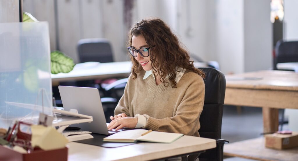 Young happy professional business woman employee sitting at desk working on laptop in modern corporate office interior. Smiling female worker using computer technology typing browsing web.