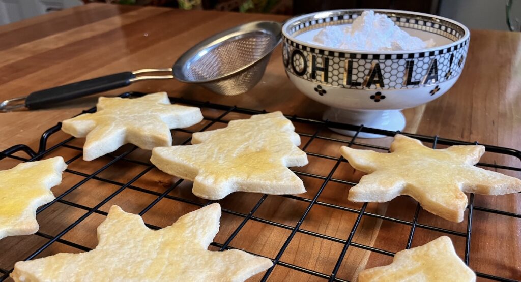 Cutout Christmas cookies on cooling rack with bowl of powdered sugar