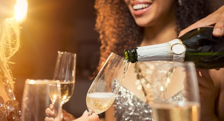 Hand pouring champagne from bottle into glasses with friends around him. Closeup of hand pouring white wine in flutes during party. Detail shot of new year’s eve celebration.