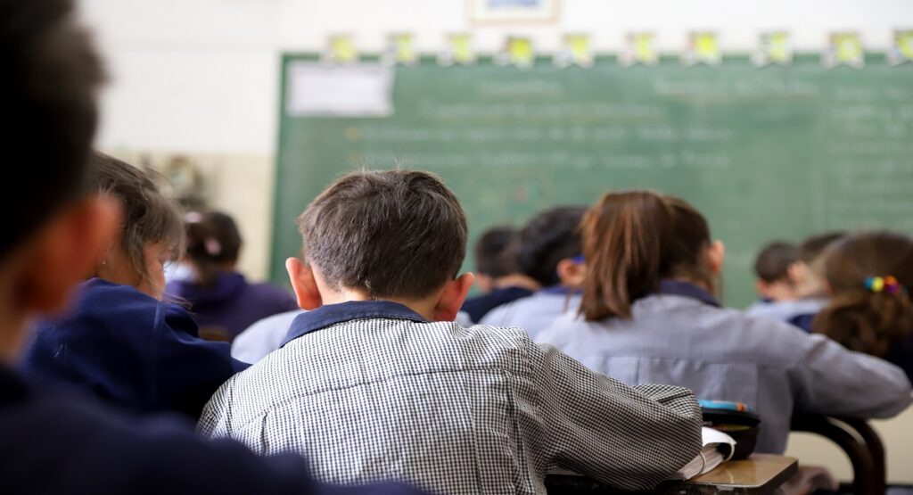 A group of children sit in desks in a classroom, facing a blackboard