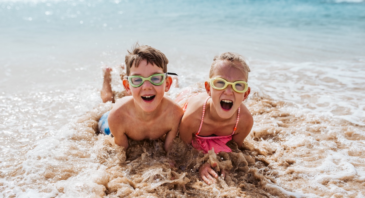 Siblings playing on beach, lying in water, having fun. Smilling girl and boy in swimsuits, swimming googles on sandy beach of Canary islands. Concept of family beach summer vacation with kids.