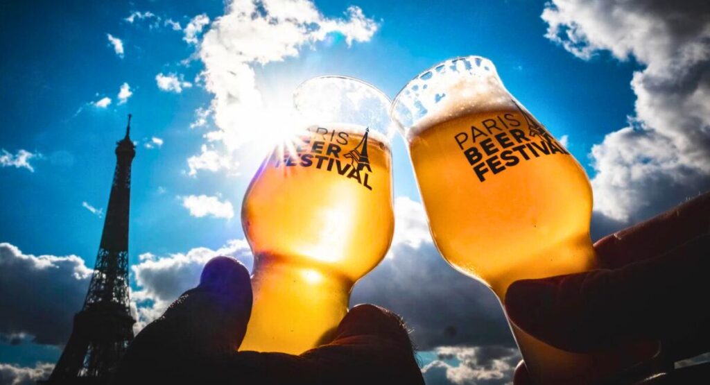 Two people raising beer glasses against a blue sky with Eiffel Tower in the background.