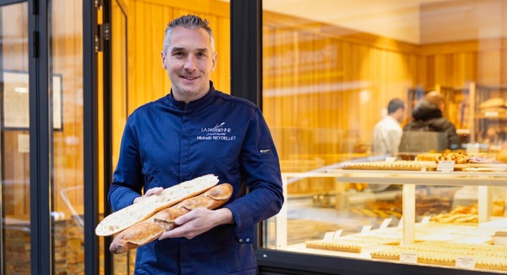 Baker holding baguettes while standing in front of window display