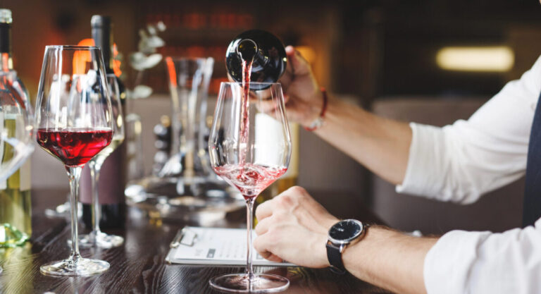 A sommelier pours red wine into stem glass at table.