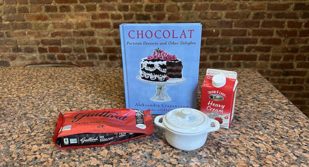 Blue book with illustration of chocolate cake, next to heavy cream carton, cocotte, and bag of chocolate chips