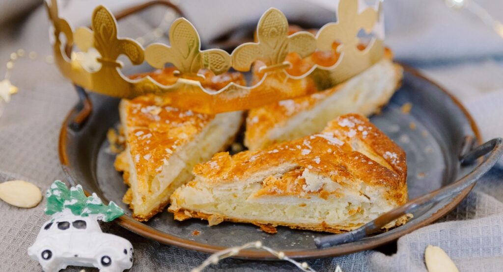 In a retro metal plate delicious three pieces of royal galette with filling on a gray kitchen napkin on a table with a golden crown, a toy and a glowing garland, side view close-up. Concept time holiday, time to eat galette.
