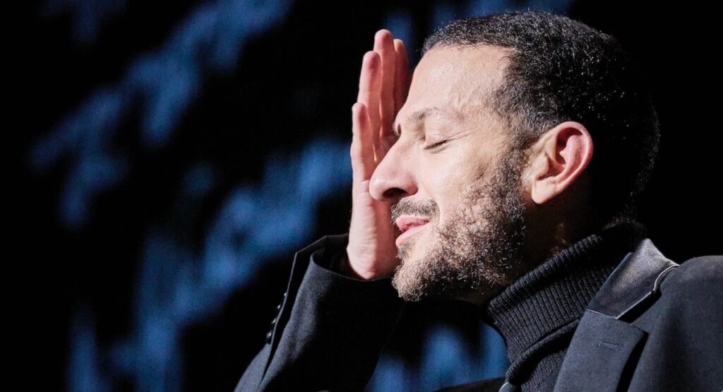 Man in black turtleneck putting a hand to his face.