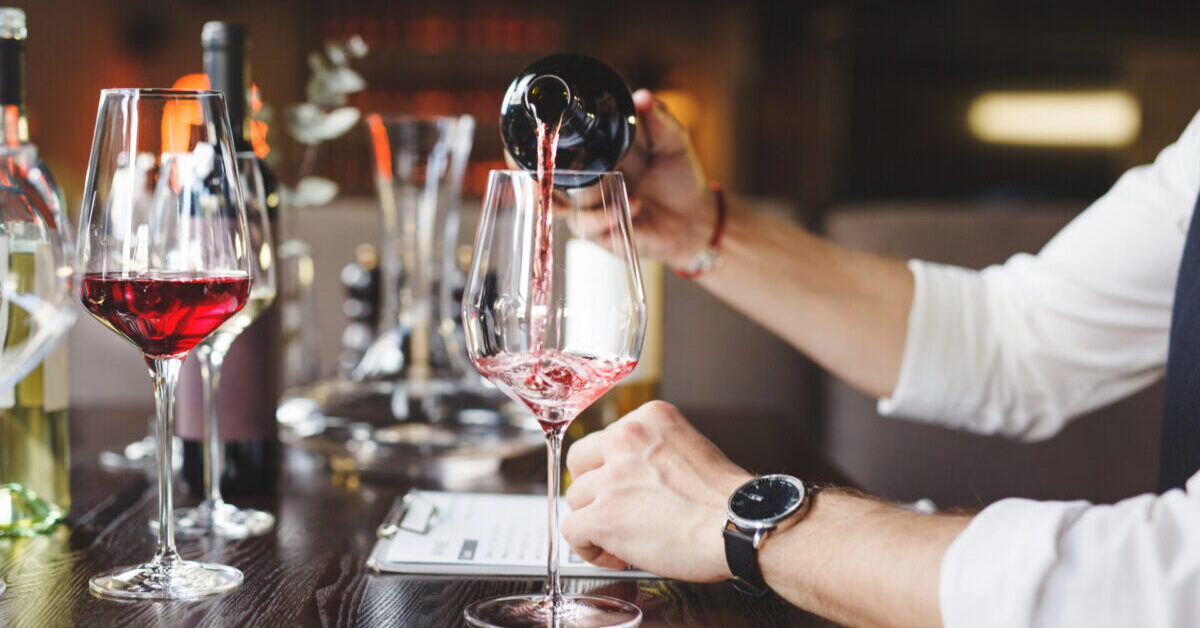 A sommelier pours red wine into stem glass at table.