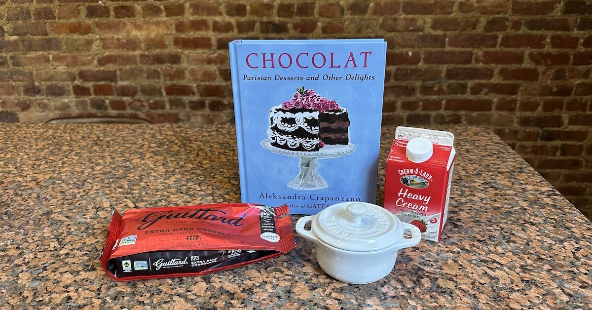 Blue book with illustration of chocolate cake, next to heavy cream carton, cocotte, and bag of chocolate chips