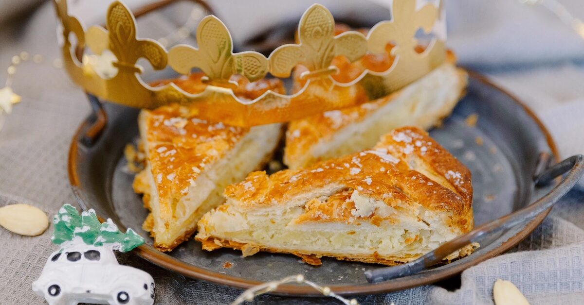 In a retro metal plate delicious three pieces of royal galette with filling on a gray kitchen napkin on a table with a golden crown, a toy and a glowing garland, side view close-up. Concept time holiday, time to eat galette.