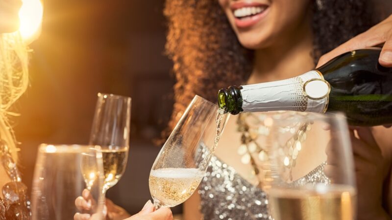 Hand pouring champagne from bottle into glasses with friends around him. Closeup of hand pouring white wine in flutes during party. Detail shot of new year’s eve celebration.