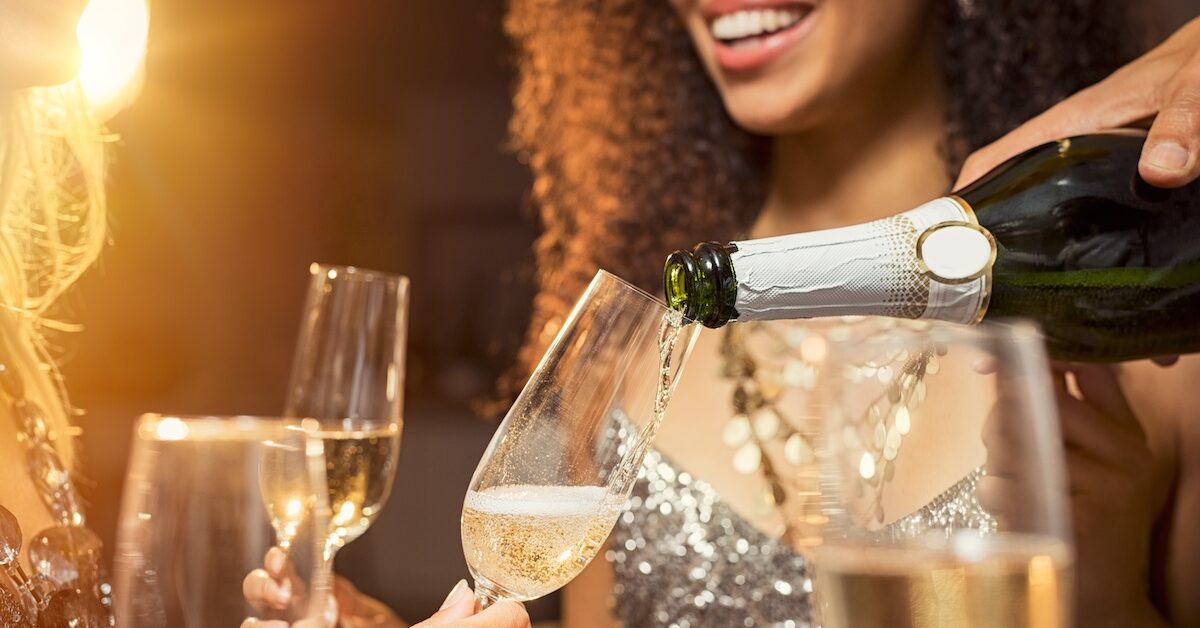 Hand pouring champagne from bottle into glasses with friends around him. Closeup of hand pouring white wine in flutes during party. Detail shot of new year’s eve celebration.