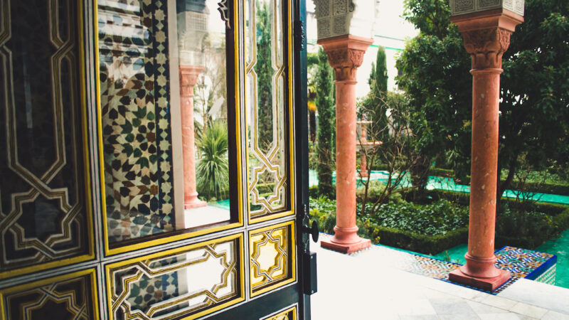 Ornamental glass door with golden geometric design facing out towards Oriental courtyard with marble pillars, garden, and emerald tiled floor. Grand Mosque of Paris.