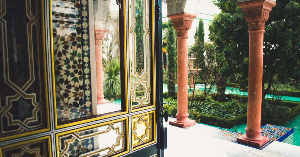 Ornamental glass door with golden geometric design facing out towards Oriental courtyard with marble pillars, garden, and emerald tiled floor. Grand Mosque of Paris.