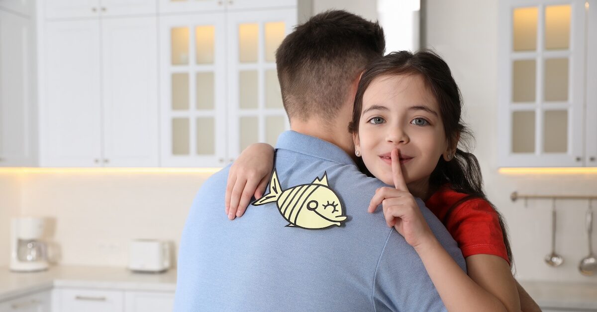 Cute little girl sticking paper fish to father's back at home